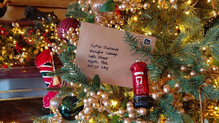 A Christmas tree decorated with warm white lights, gold berry garlands and colourful baubles, featuring a handwritten letter addressed to Father Christmas nestled among the branches, with a festive fireplace and Christmas stocking in the background.